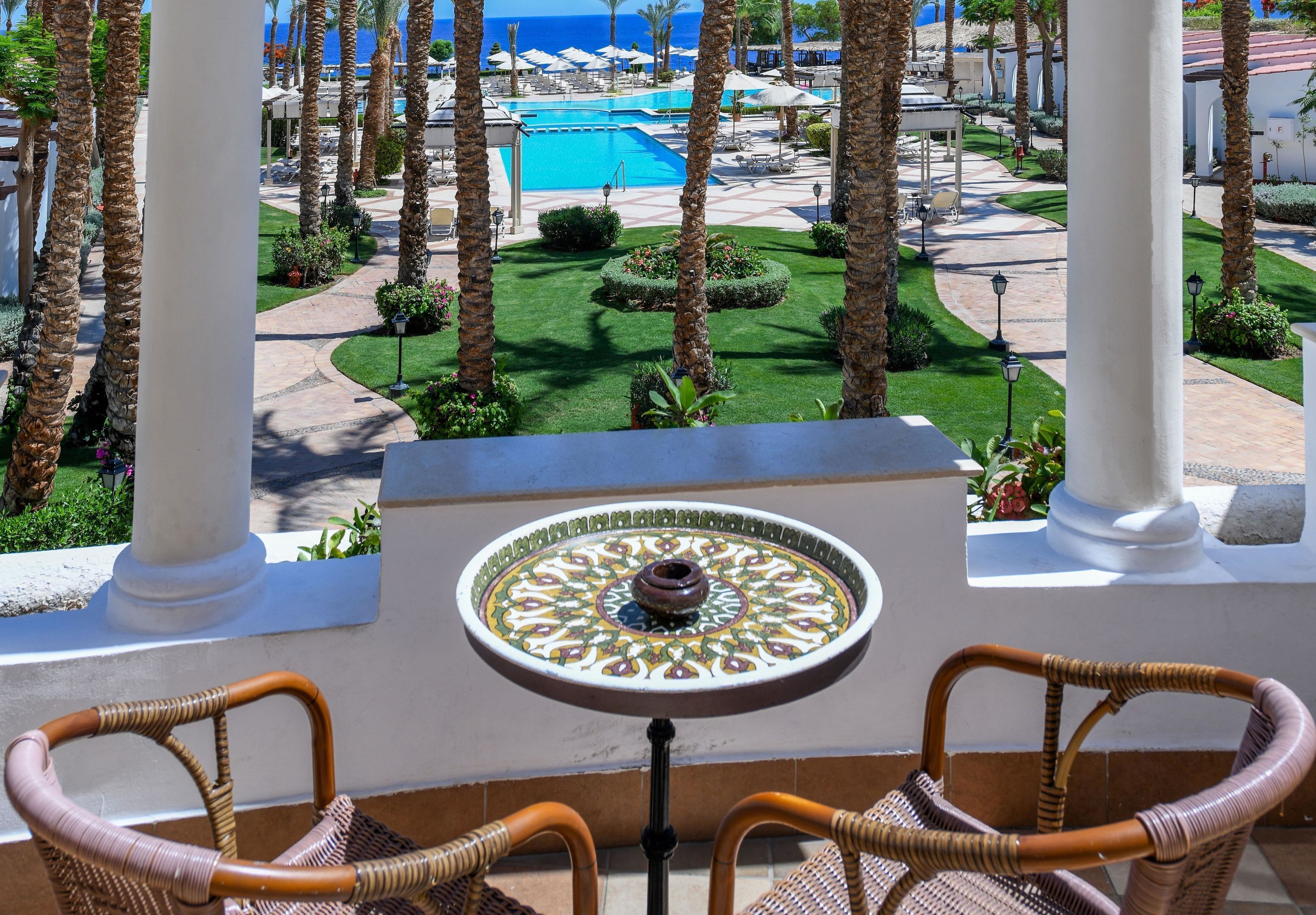 Un balcon avec des fauteuils en osier, une table décorative et une vue sur une piscine et des palmiers.