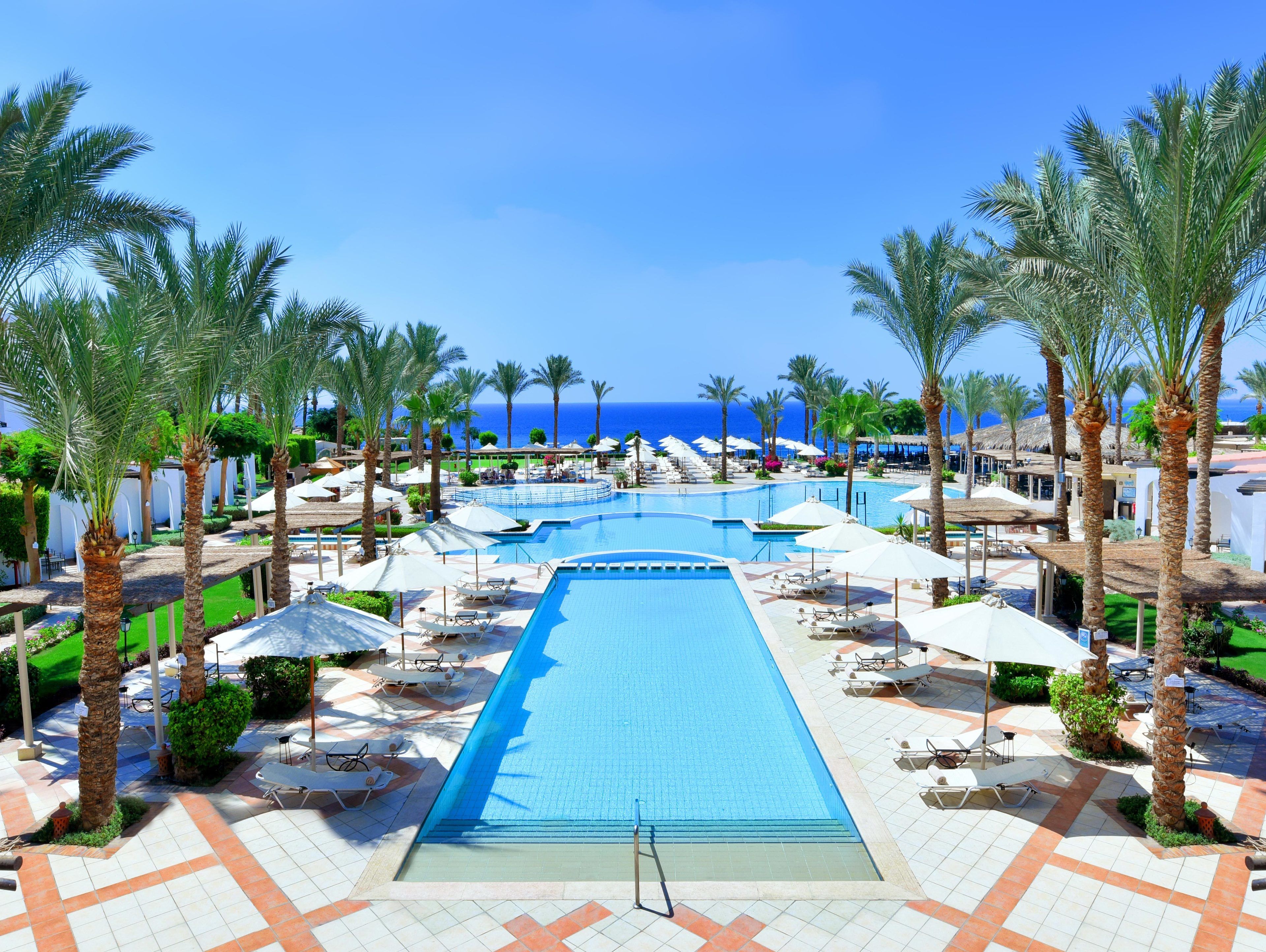 A resort pool area with multiple swimming pools, sun loungers, and palm trees.