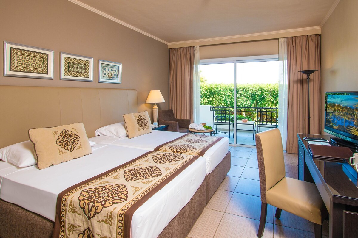 A hotel room with a large bed, a desk with a computer, a chair, a TV, and a balcony with a view of greenery.