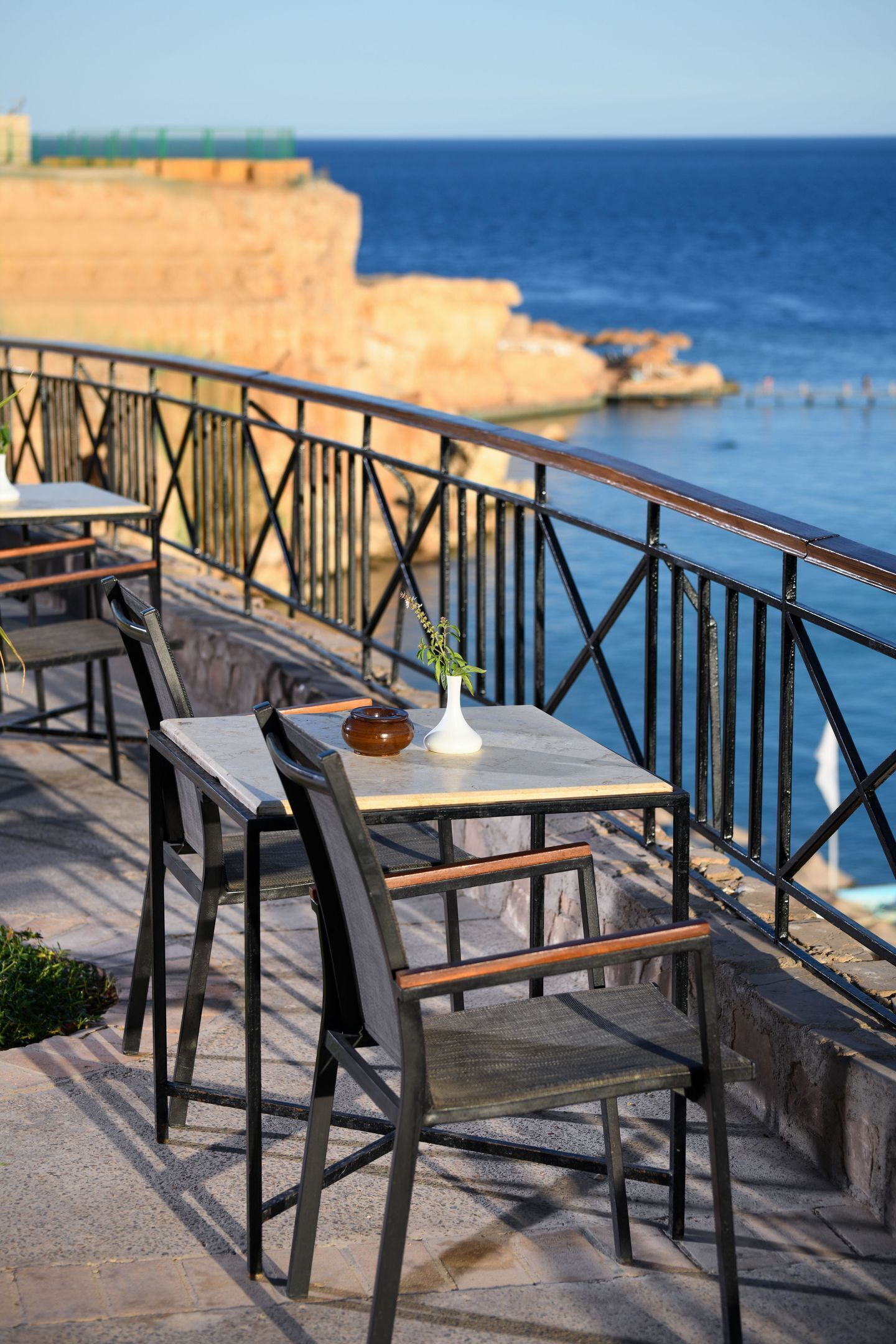 An outdoor seating area with tables and chairs, a railing, and a view of the sea.