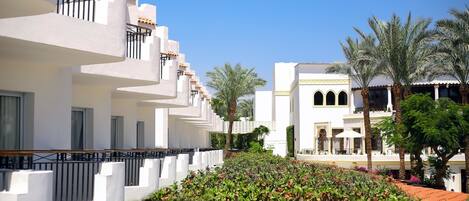 A hotel with white exterior walls, balconies, and a well-maintained garden with shrubs and palm trees.