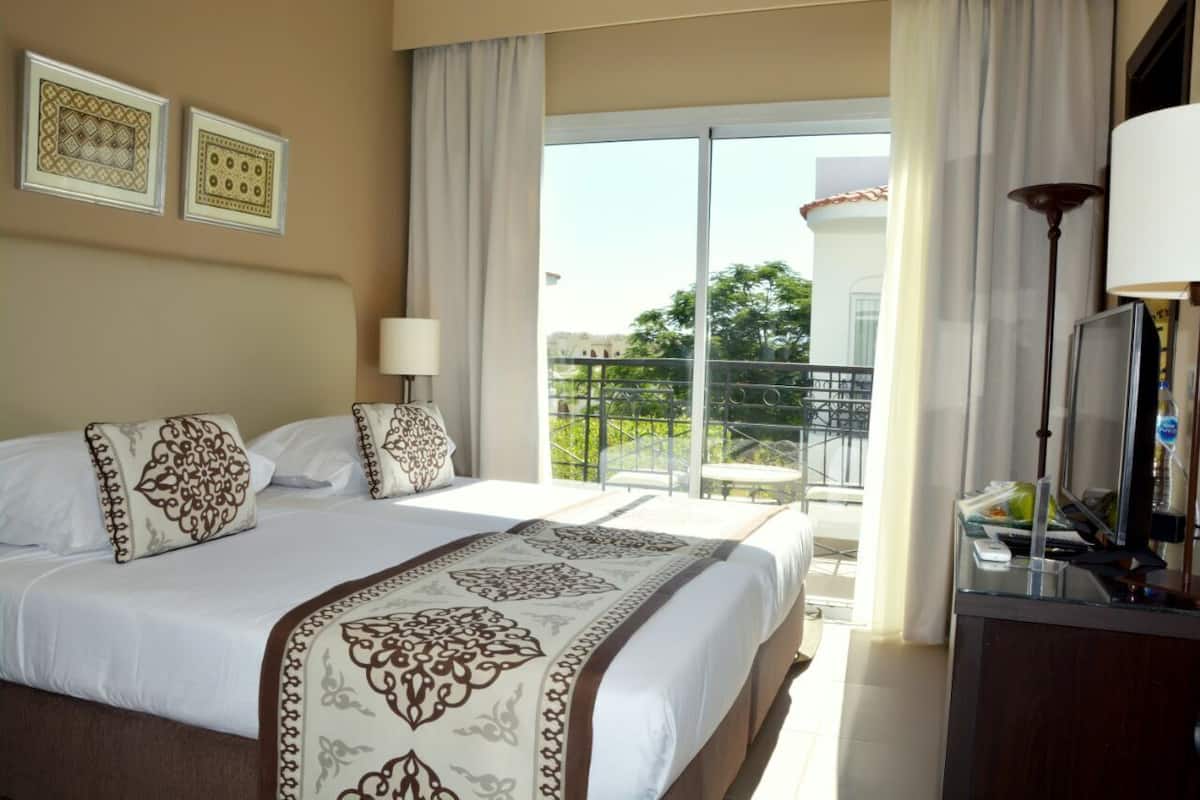 A hotel room with a large bed, two bedside lamps, a desk with a lamp, and a view of a balcony with a railing.