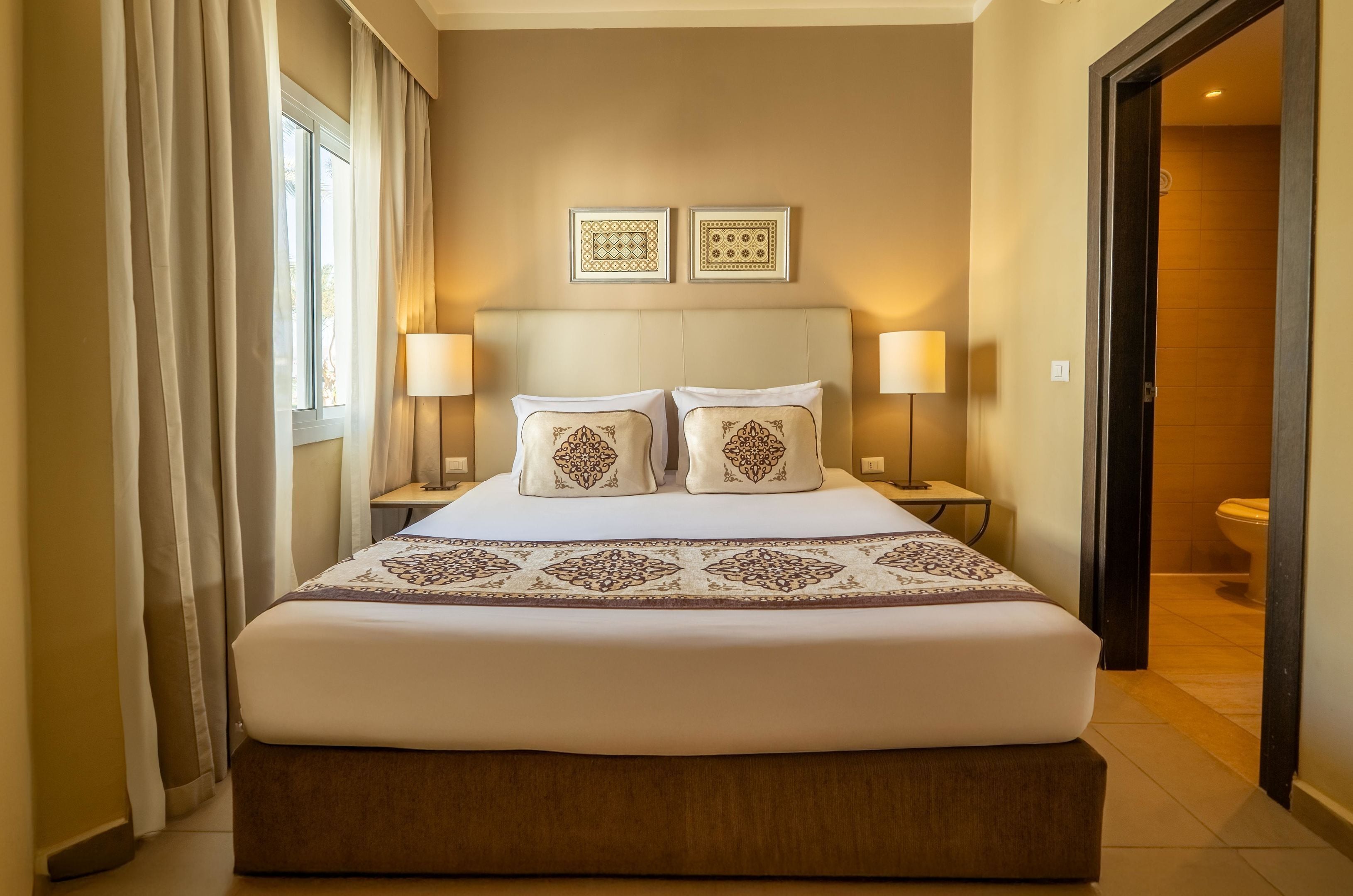 Una camera d'albergo con un letto grande, due comodini con lampade, una finestra con tende e due quadri appesi al muro.