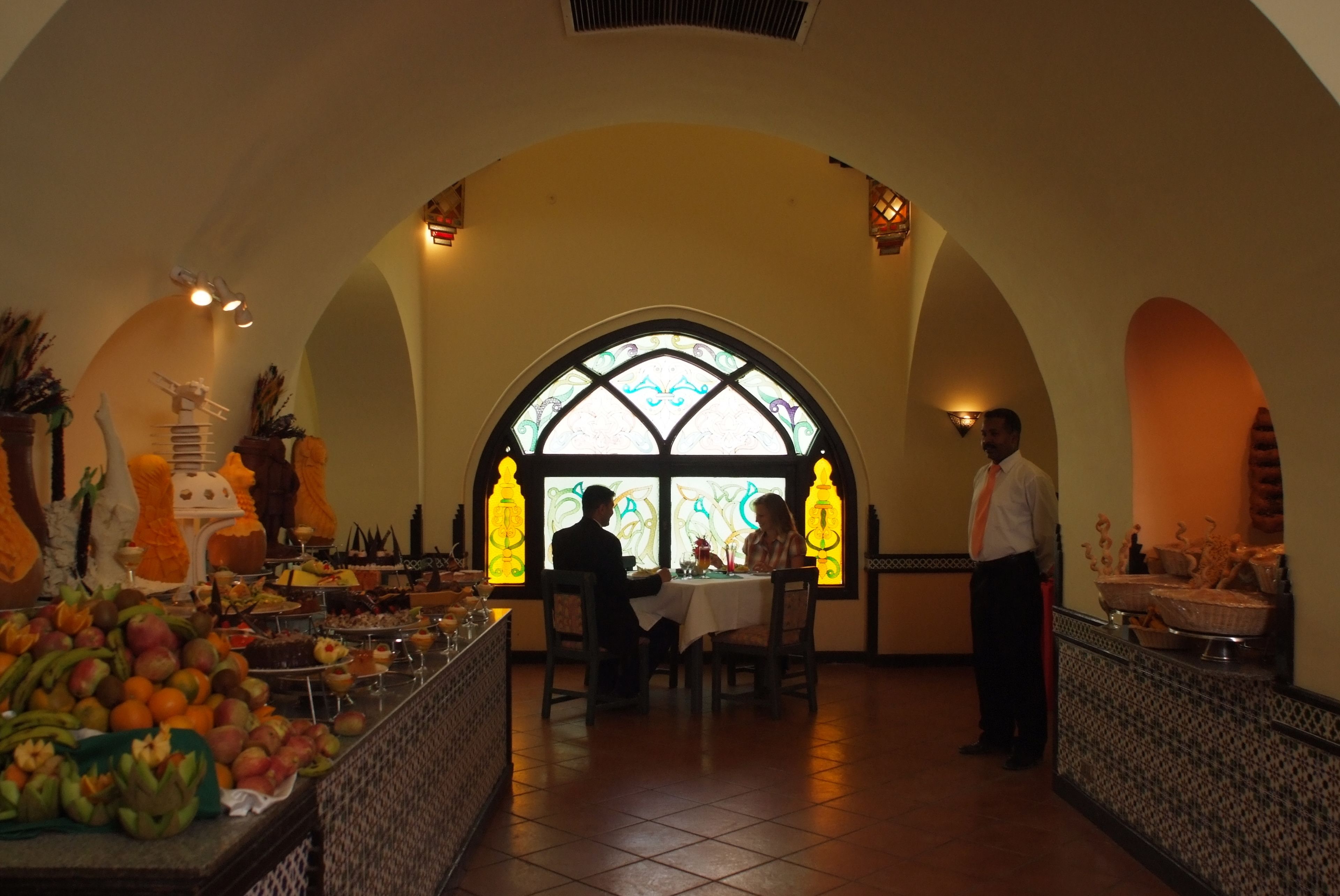 Een restaurant met een hoog gewelfd plafond, een centraal geplaatst glas-in-loodraam en een buffetgedeelte met verschillende soorten fruit en gerechten.