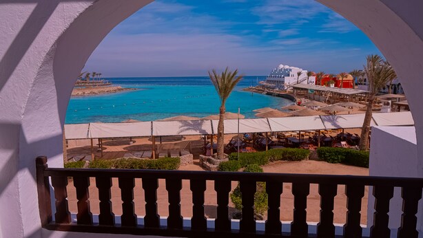 Une vue sur la plage depuis un balcon doté d’une balustrade, un espace piscine avec des parasols, et un bâtiment à la façade blanche.