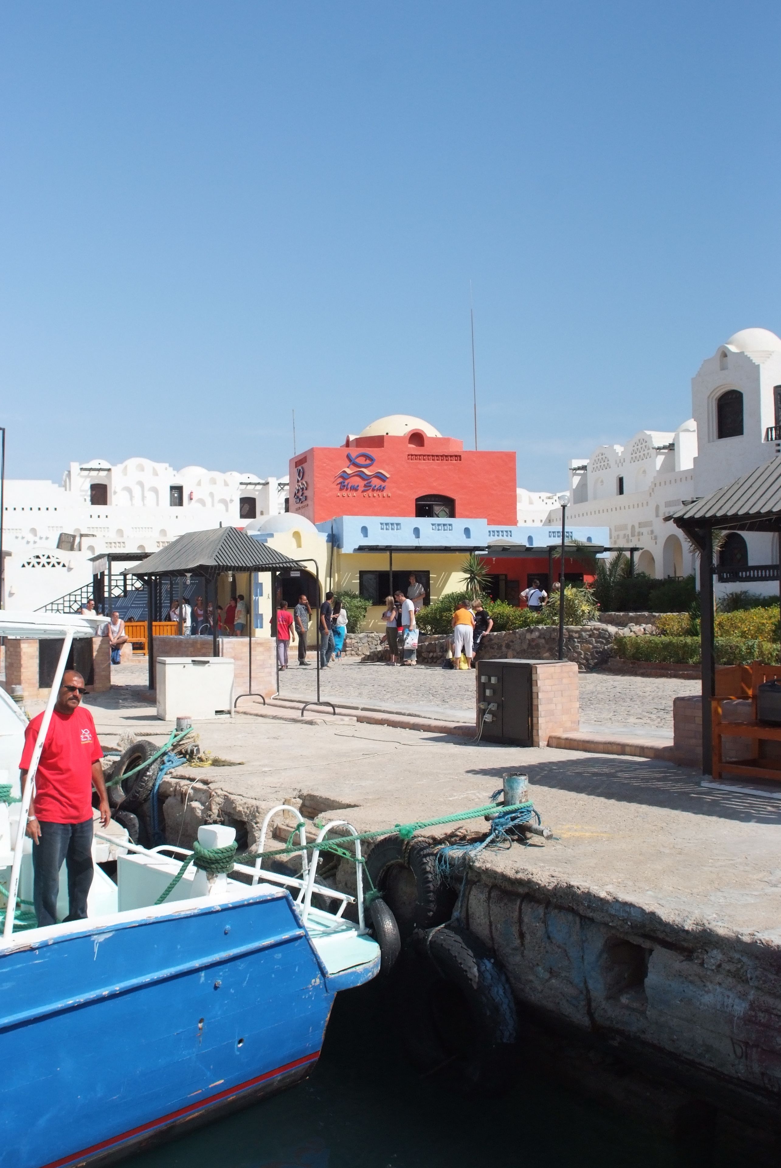 A coastal area with a blue boat docked at a pier, a building with a red and yellow facade, and a clear blue sky.