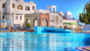 Une piscine à l’eau bleue, entourée de bâtiments blancs ornés d’arches et de coupoles, et de chaises longues bleues.