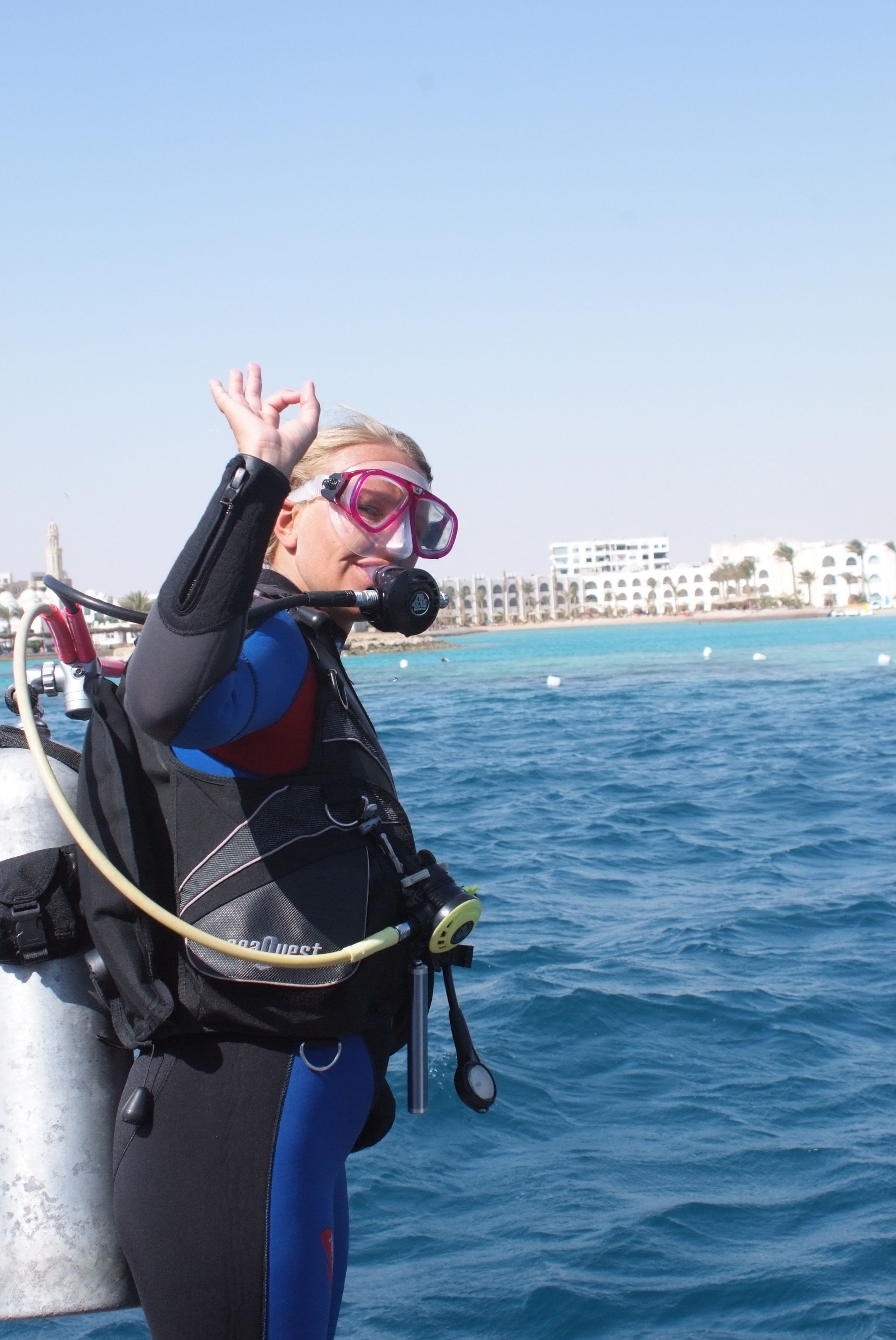 A diver in a blue and black wetsuit with a scuba tank and regulator, standing by a boat with a clear blue sea and a coastal building in the background.