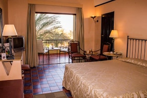 A hotel room with a bed, a desk with a TV, a balcony with a table and chairs, and a view of palm trees. - Solymar Makadi Sun - All inclusive (Makadi Bay)
