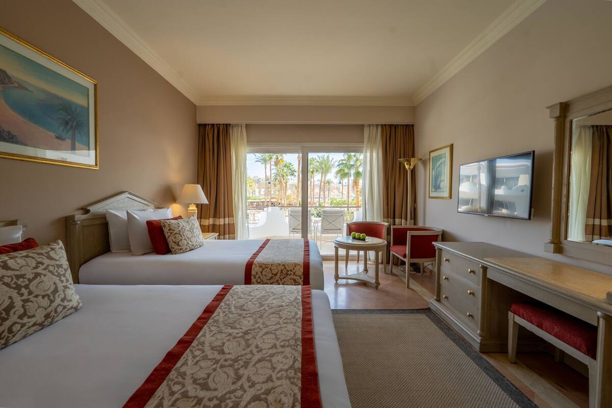 Een hotelkamer met twee bedden, een bureau, een stoel, een televisie en een balkon met palmbomen.