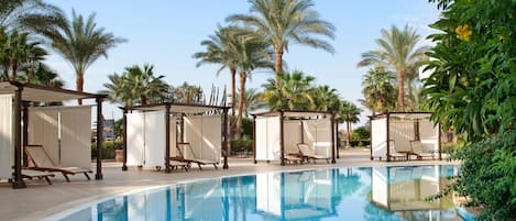 A pool area with lounge chairs and shaded seating areas, surrounded by palm trees and greenery.