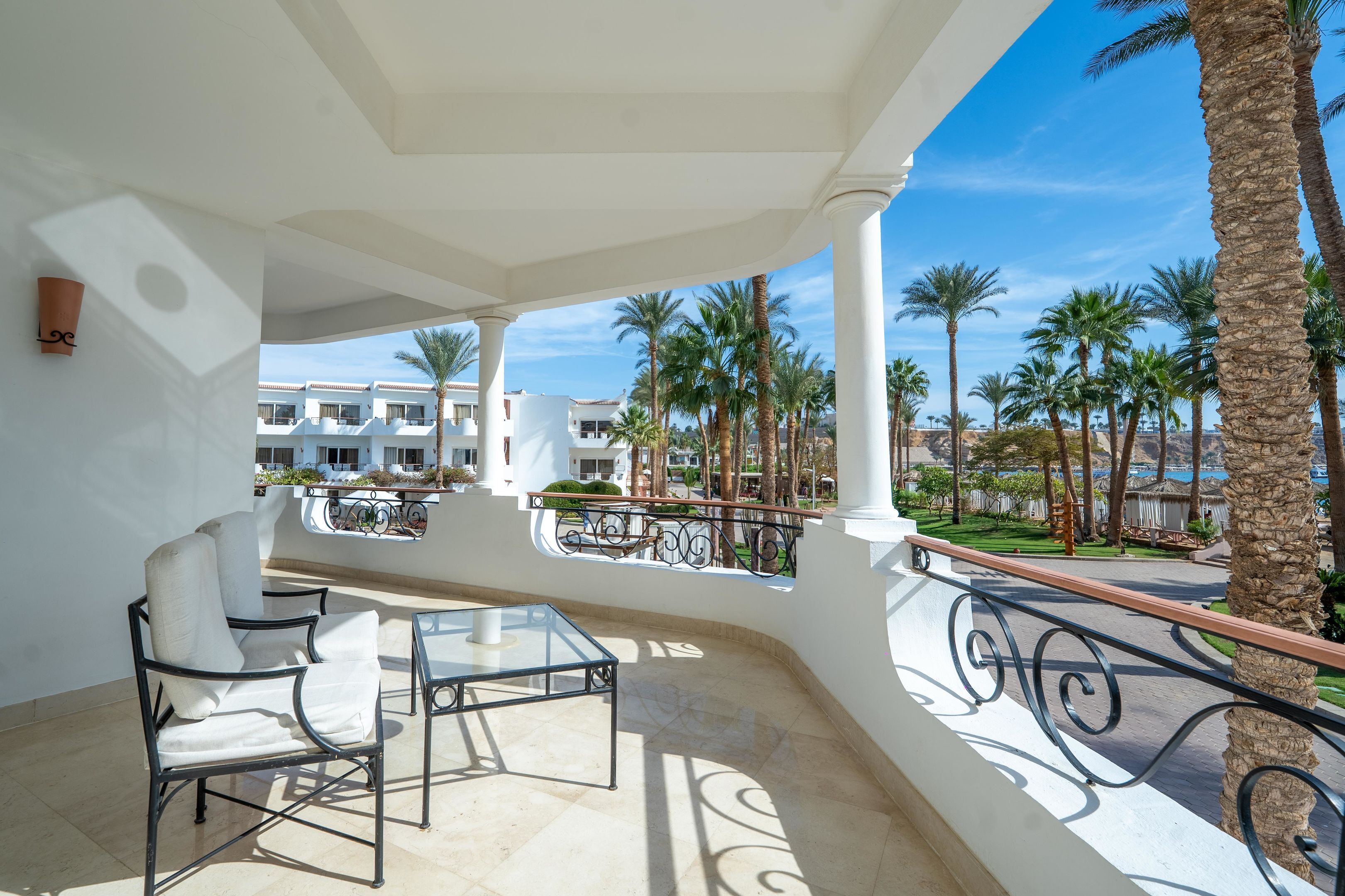 A balcony with a glass table and chairs, palm trees, and a view of a building complex.