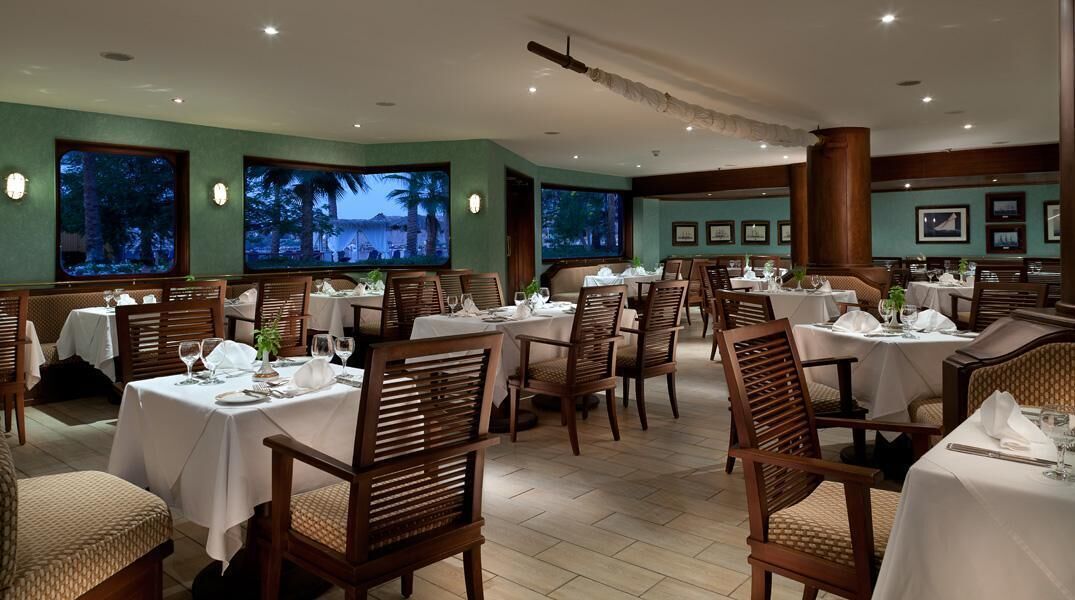 A restaurant with multiple tables and chairs, large windows with a view, and framed pictures on the walls.