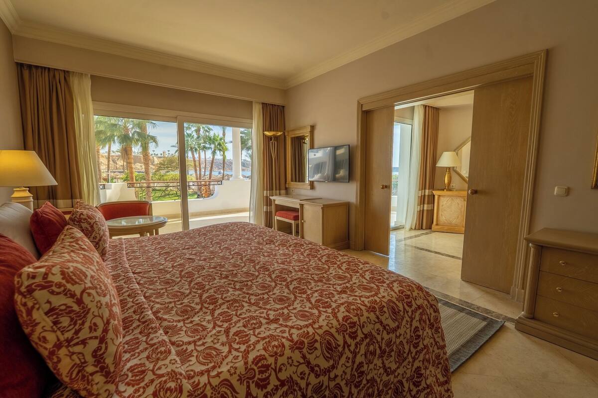 Een hotelkamer met een groot bed, een bureau met een televisie, een balkon met uitzicht op palmbomen, en een deur naar een andere kamer.