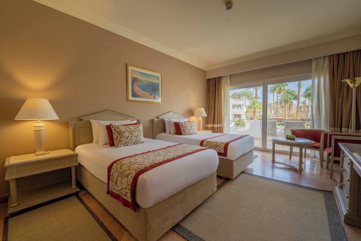 Een hotelkamer met twee bedden, een bureau, een stoel en uitzicht op palmbomen door het raam.
