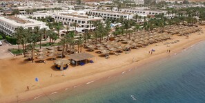 Luchtbeeld van een strandresort met rijen palmbomen, strandparasols en een groot hotelcomplex.
