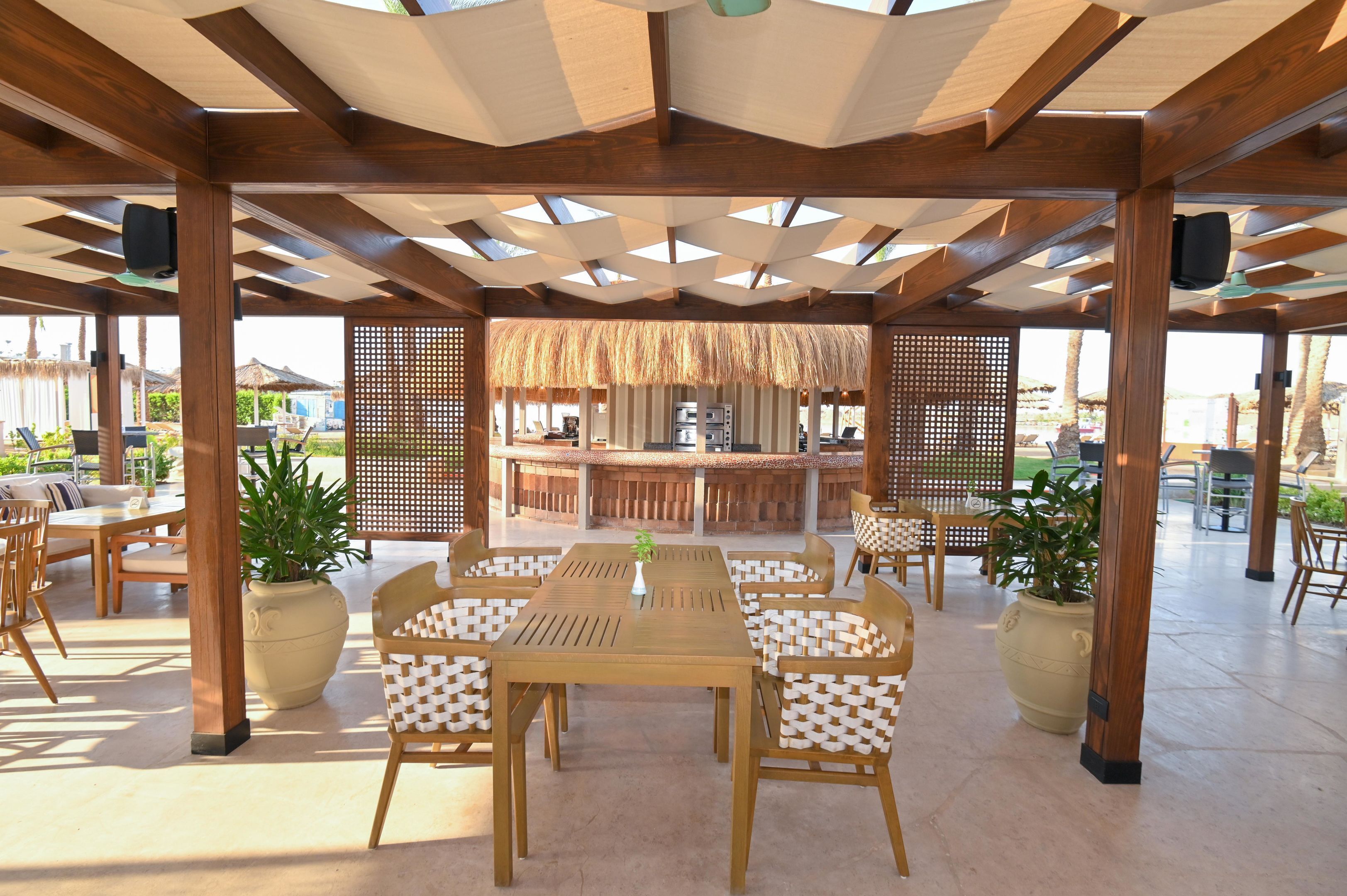 Un área de comedor al aire libre con mesas y sillas de madera, plantas en macetas y una zona de bar con techo de paja.