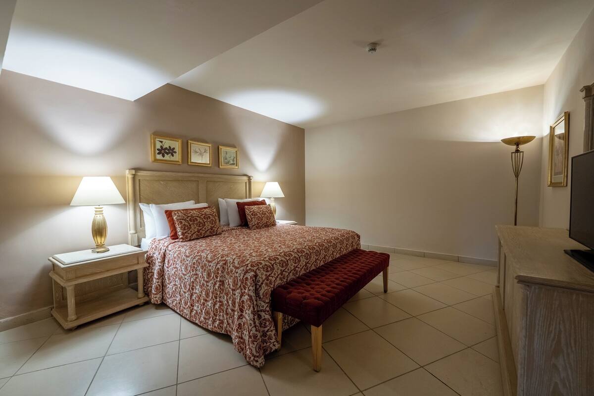 Een slaapkamer met een bed, twee nachtkastjes, een bankje, een televisie en een plafondlamp.