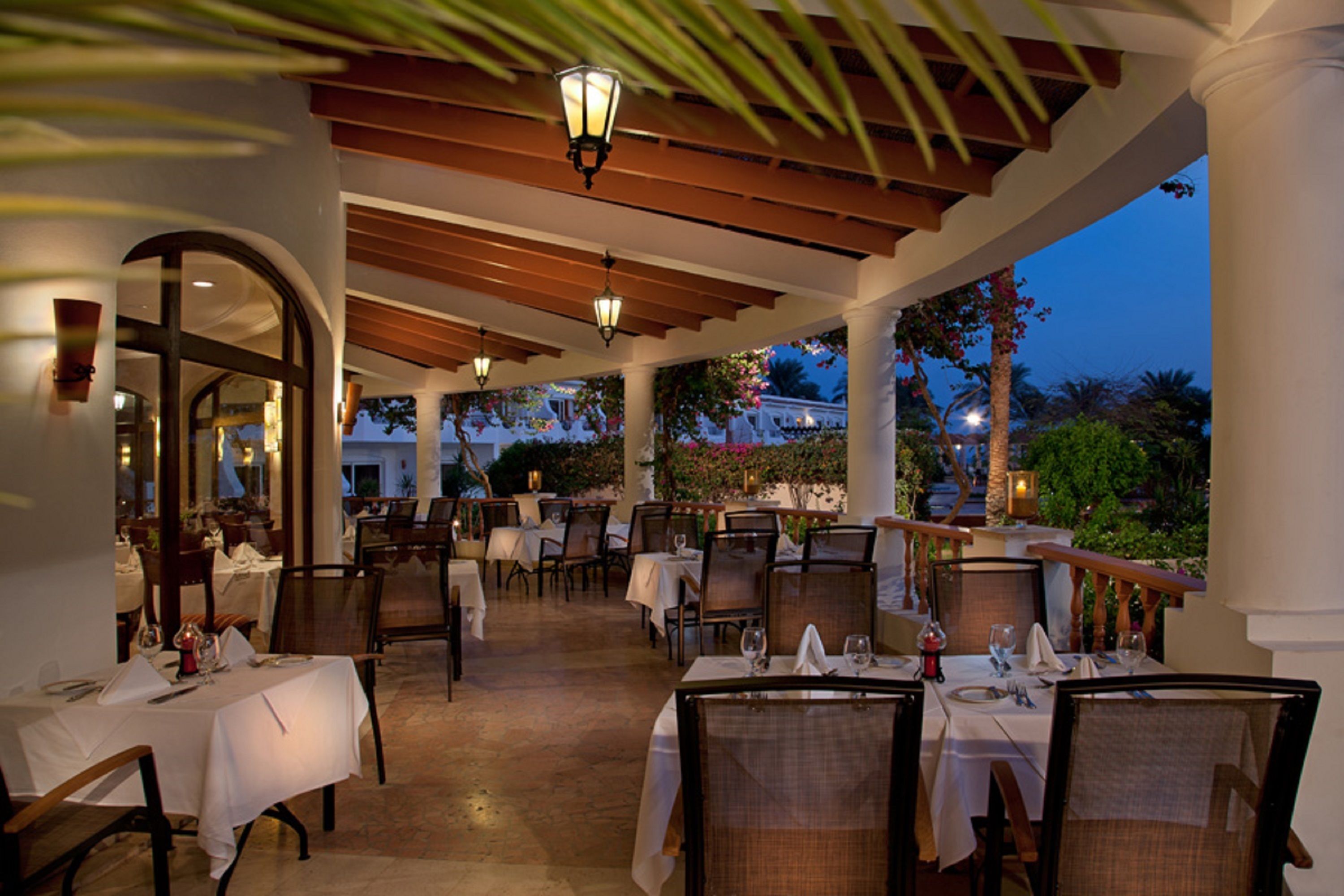 An outdoor dining area with tables set for dinner, covered by a wooden pergola with hanging lanterns, and a view of the evening sky.