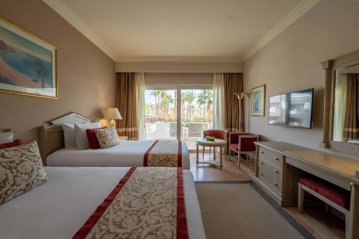 Hotelkamer met twee bedden, een bureau, een stoel, een televisie en een balkon met palmbomen.