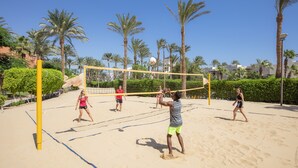 Partido de voleibol de playa en una cancha arenosa rodeada de palmeras y edificios.