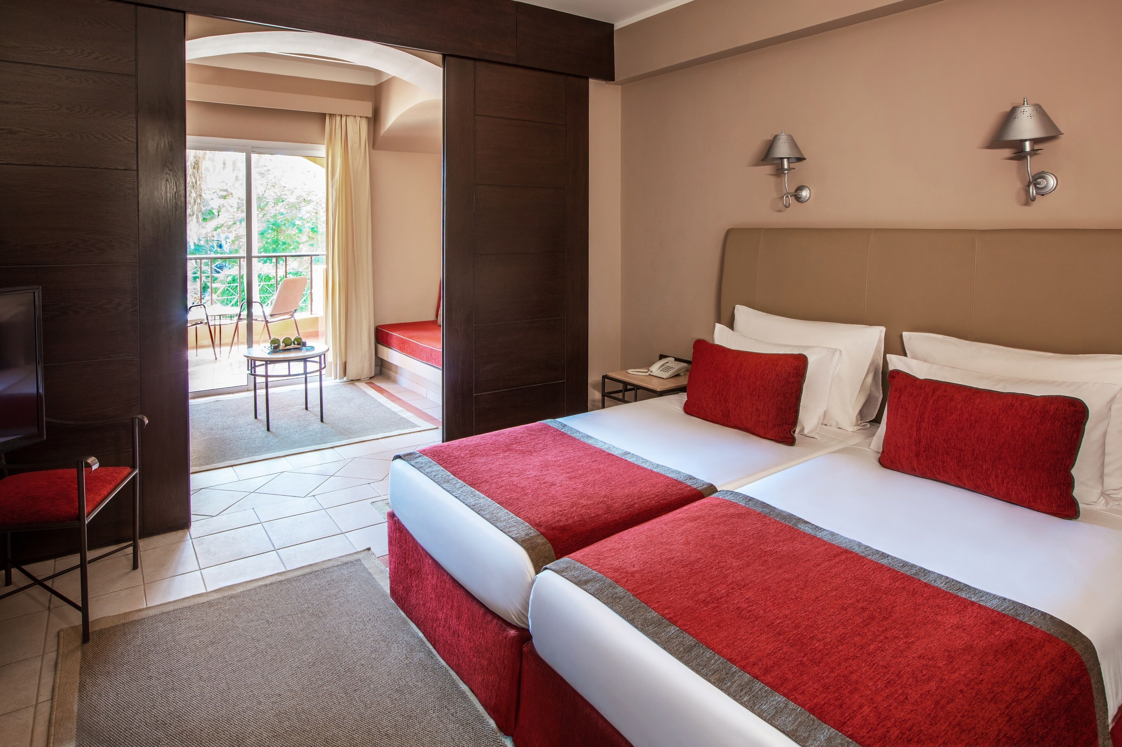 A hotel room with two beds, a TV, a balcony with a chair, and a small table.