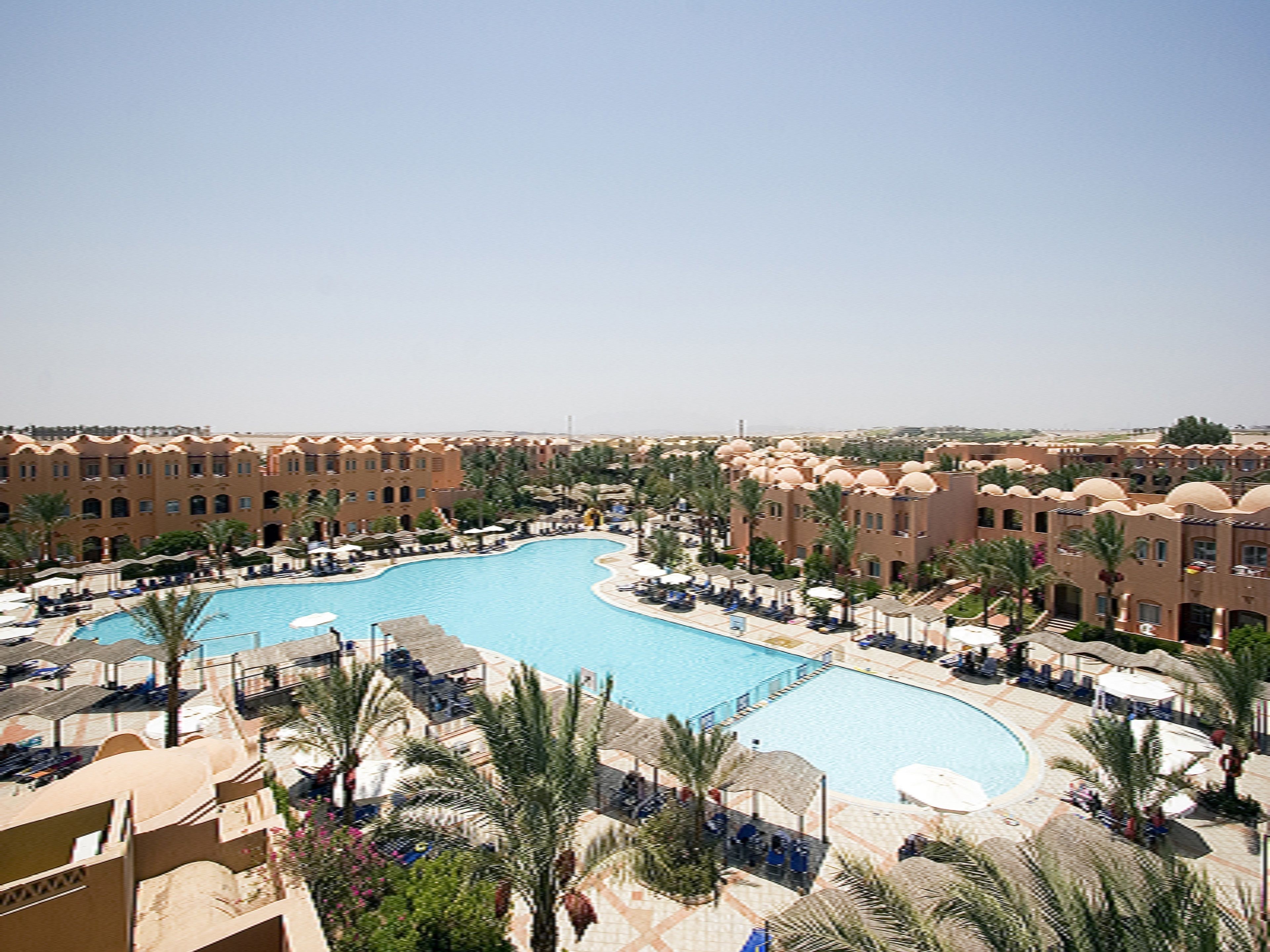 A resort with a large central pool surrounded by palm trees and sun loungers.