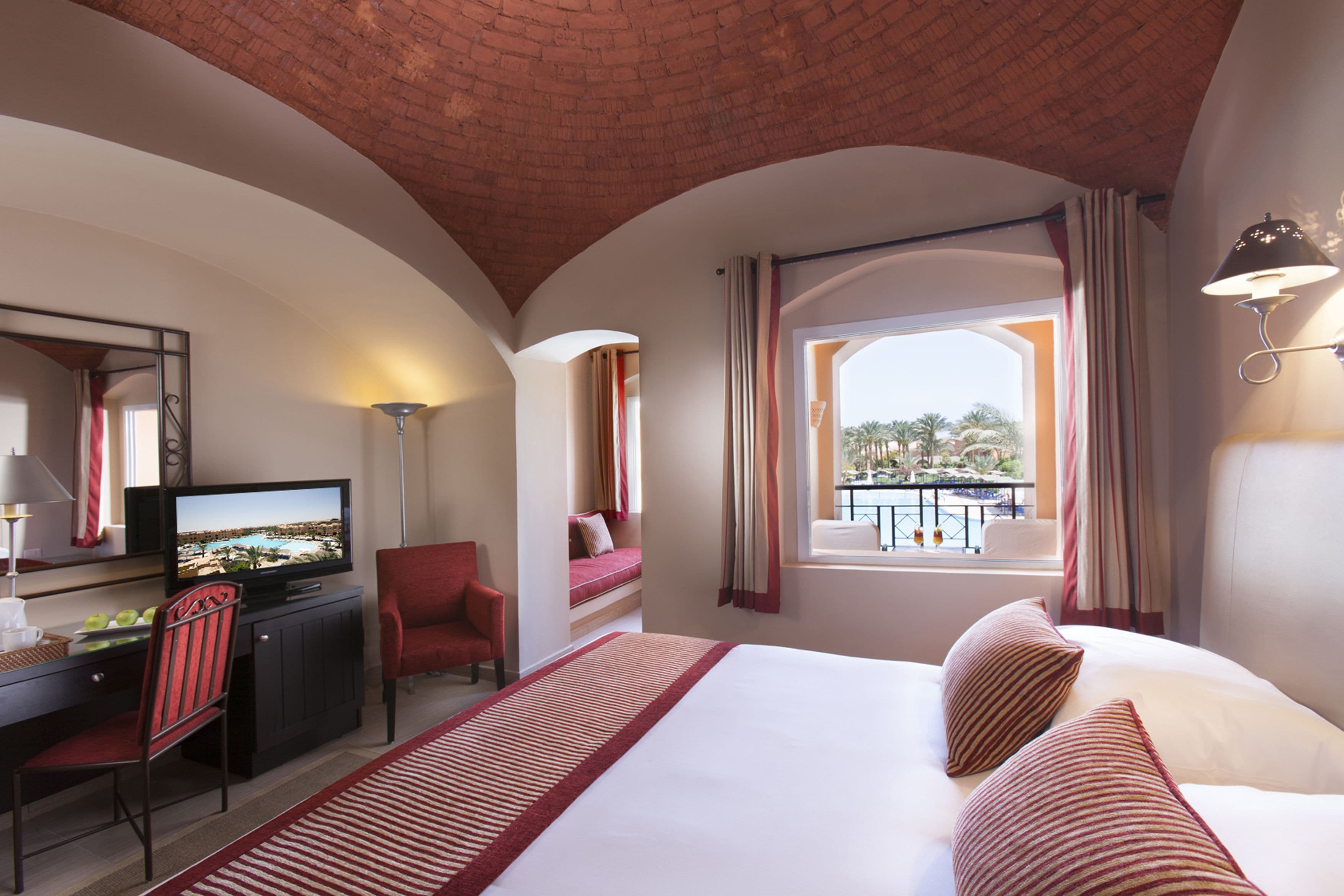 A hotel room with a bed, a desk with a TV, a chair, and a balcony with a view.