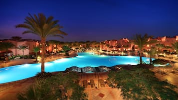 Piscina de un resort con palmeras, sillas de descanso y una iluminación nocturna.