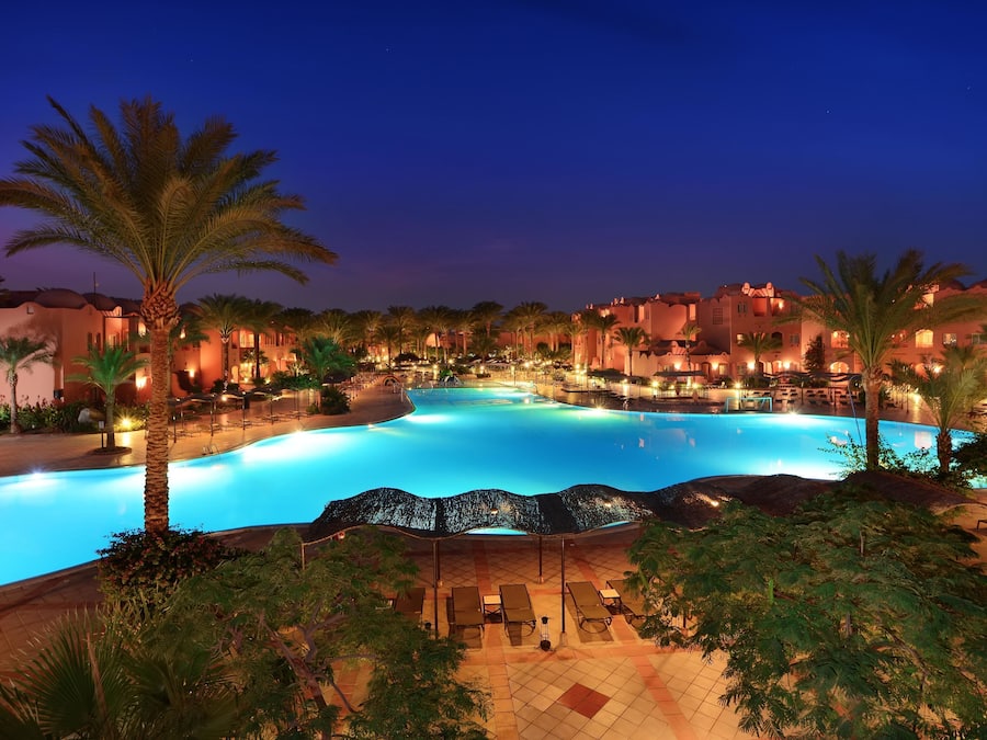 Piscina di un resort con palme, poltrone relax e illuminazione notturna.