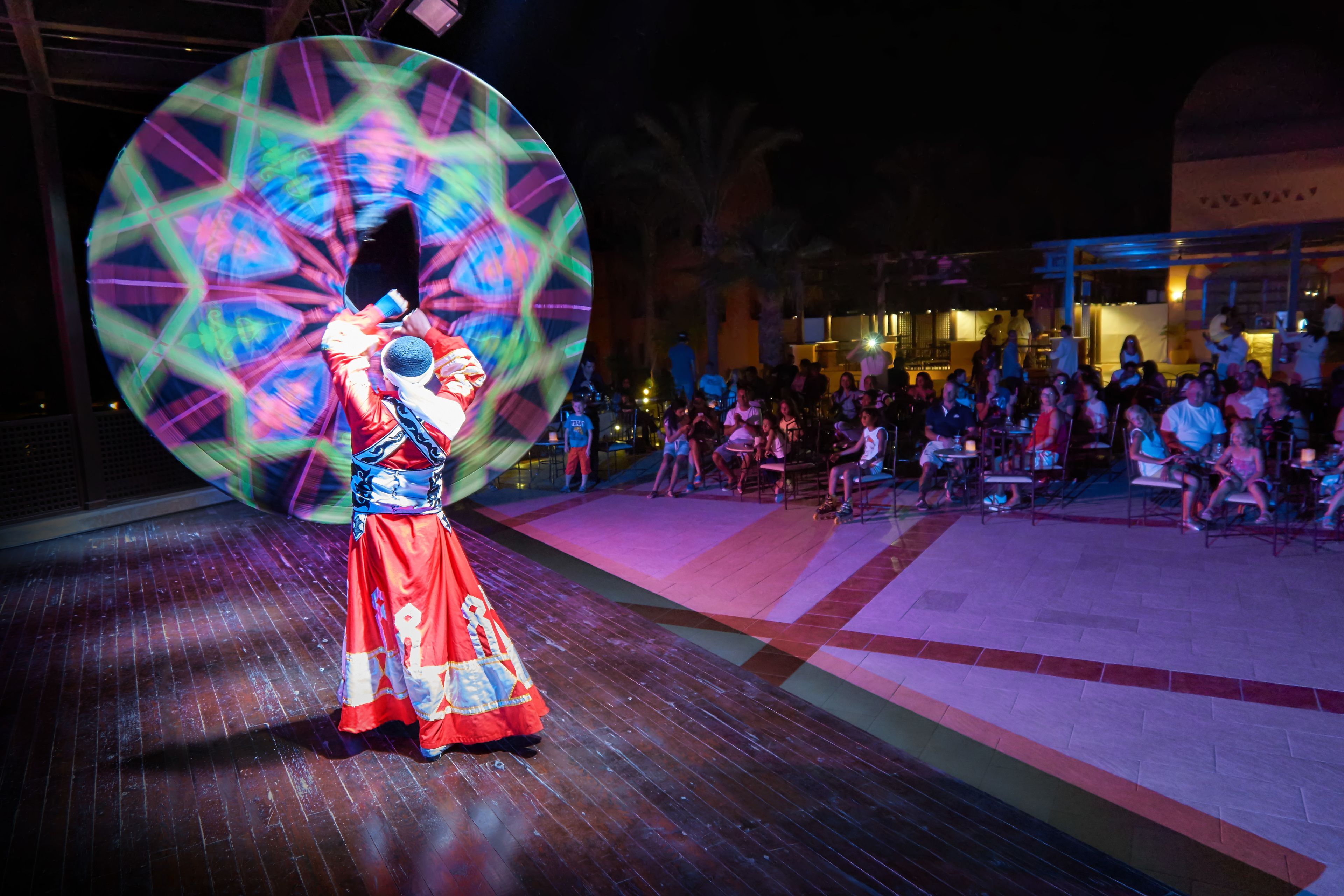 A person in traditional attire performing in front of a large, colorful circular light installation.