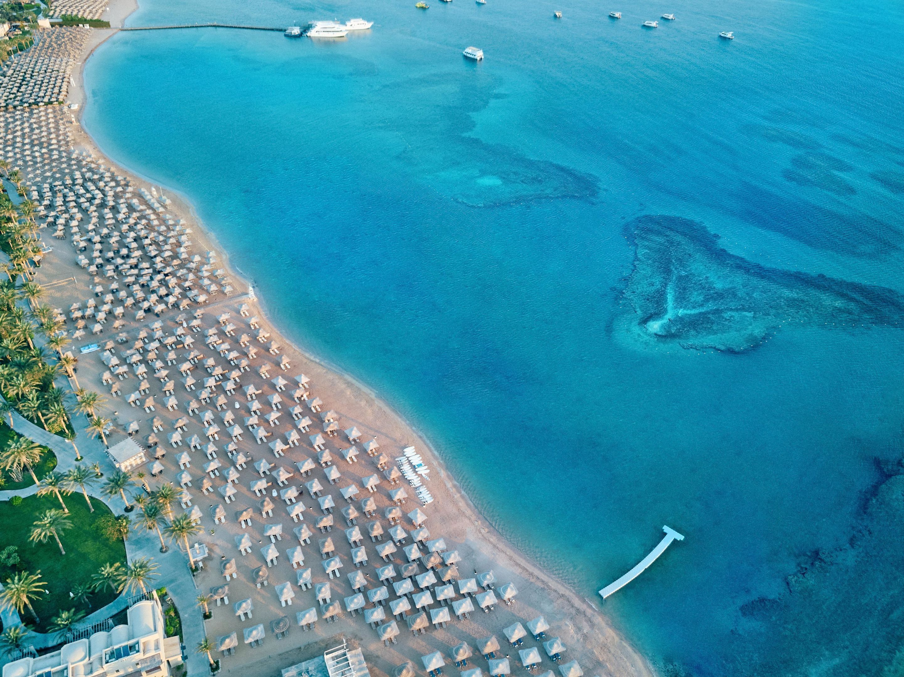 Vista aérea de una playa con numerosas sombrillas y sillas de playa, mar azul cristalino y embarcaciones en el agua.