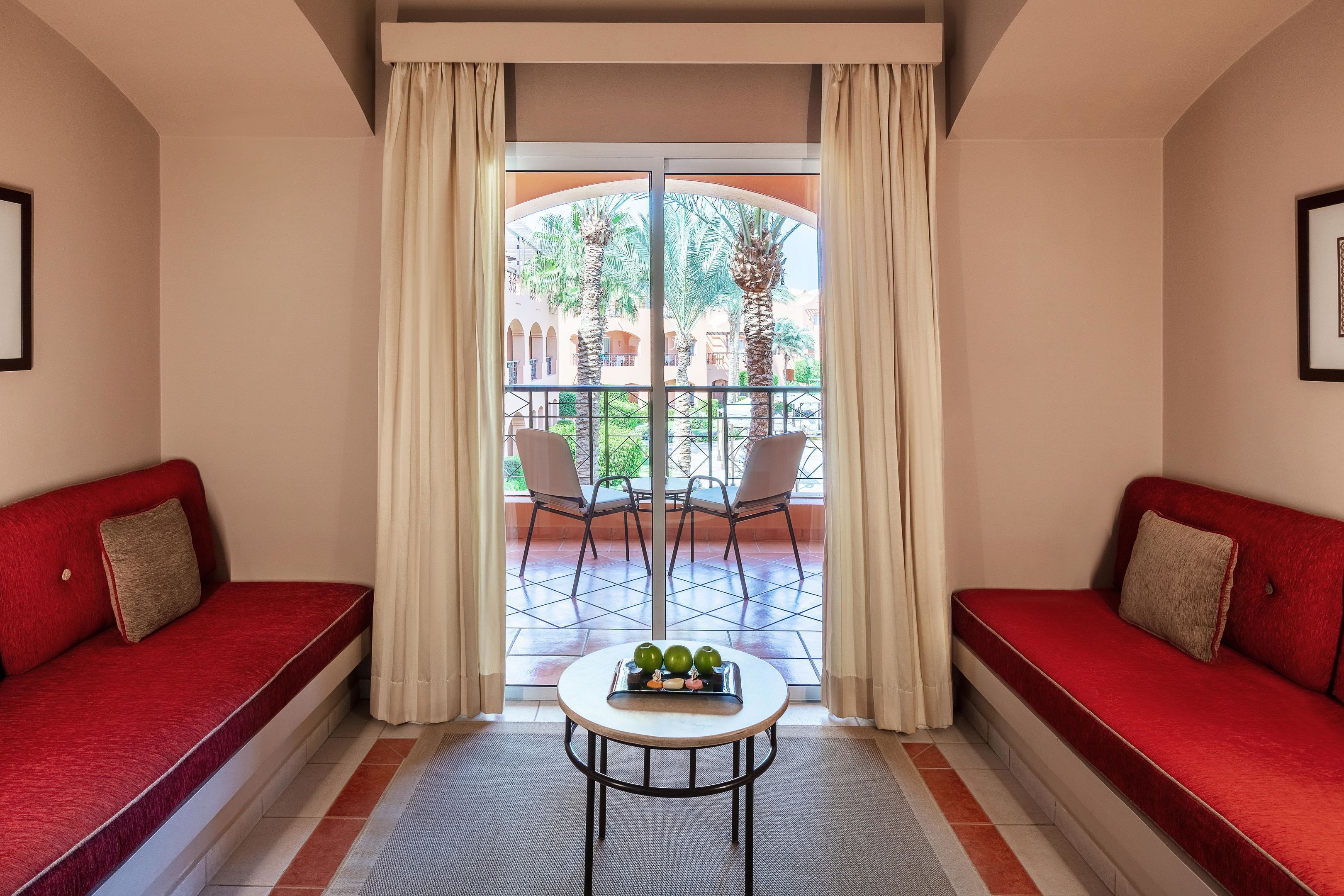 Une pièce avec un canapé rouge, une petite table ronde sur laquelle se trouve un plateau de pommes, et une vue sur une cour intérieure agrémentée de palmiers et d’un balcon.