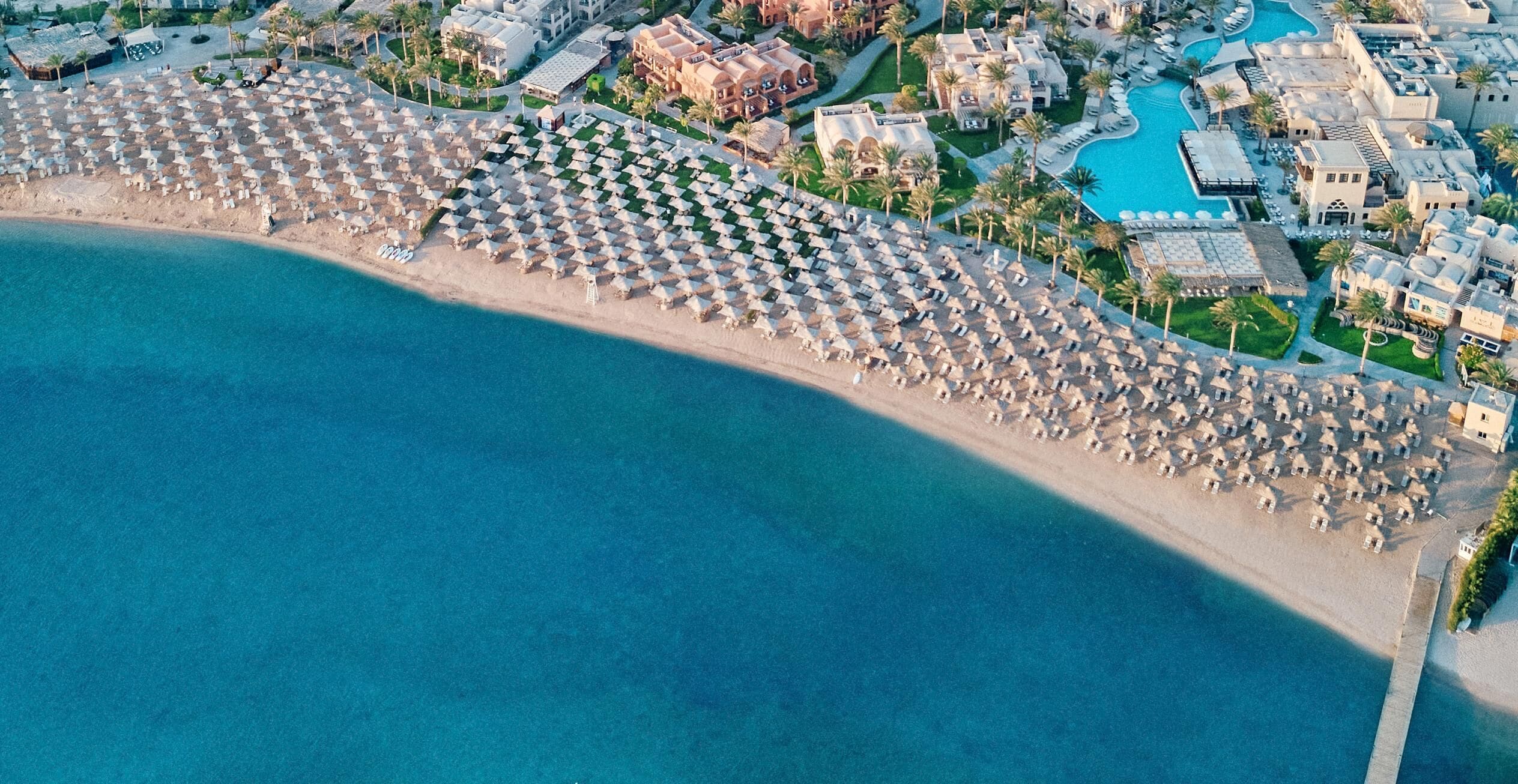 Vista aérea de un complejo turístico en la playa, con filas de sombrillas y sillas, una piscina y edificios del resort.