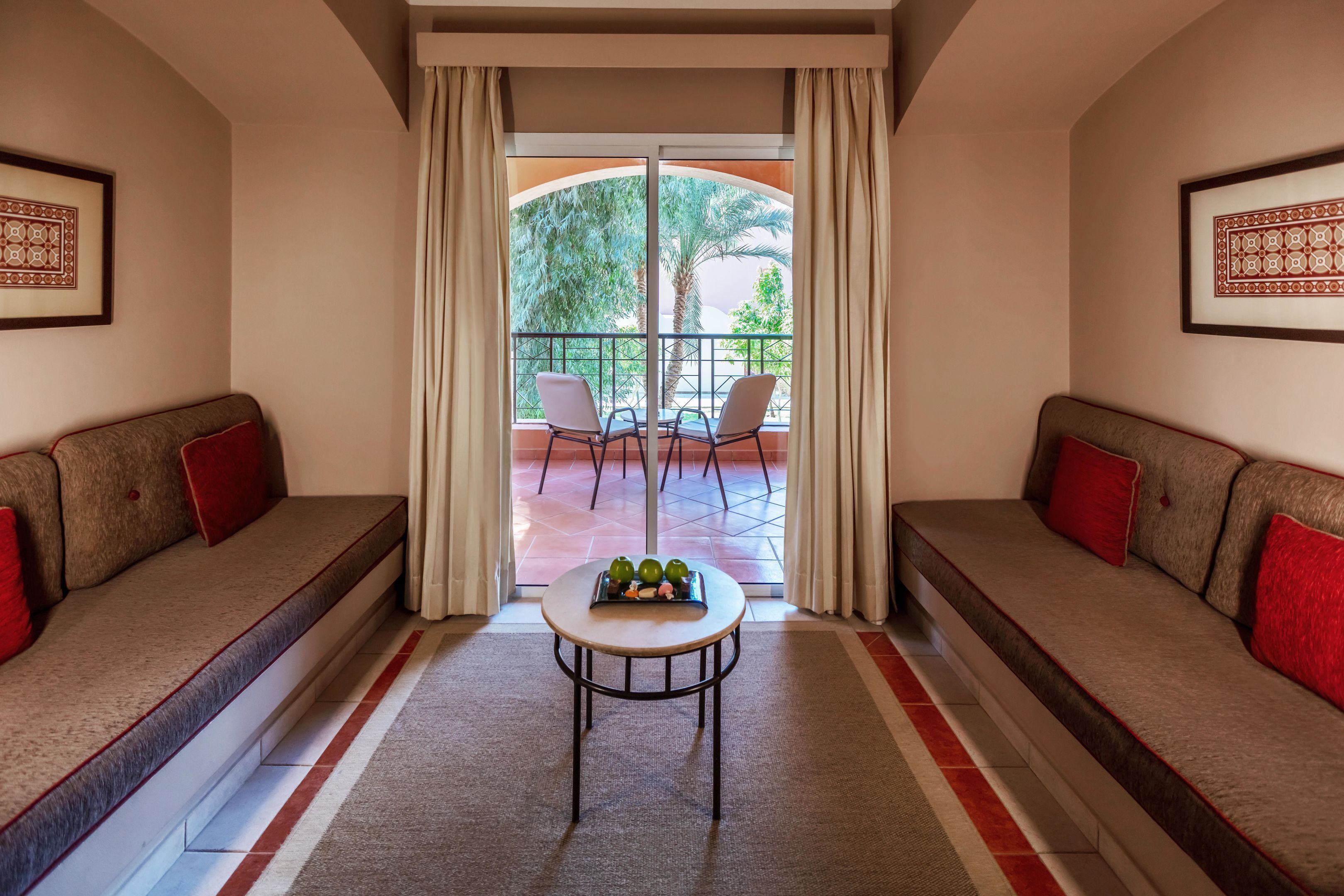 A room with a sofa, a small table with fruit, and a view of a balcony with chairs.