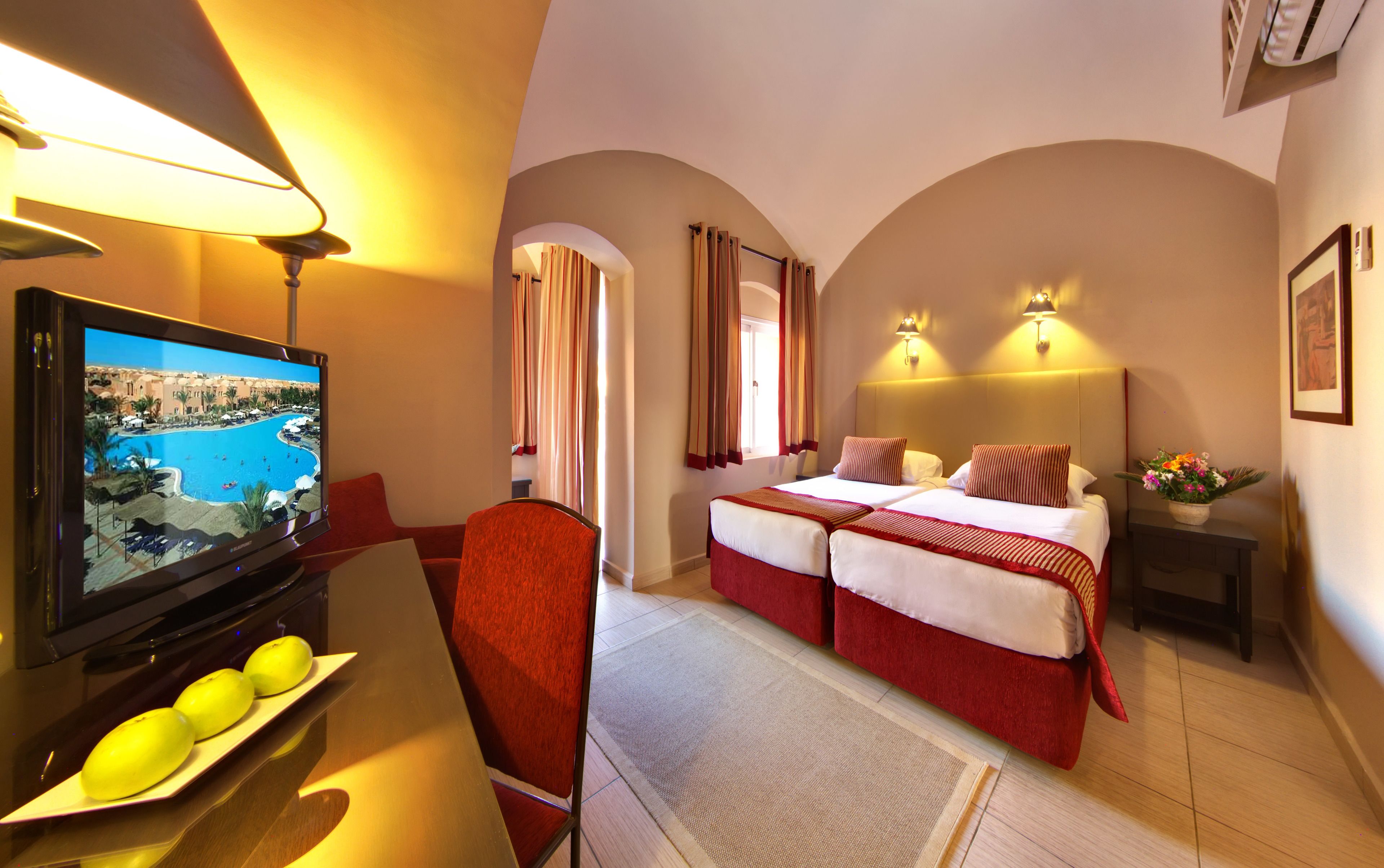 A hotel room with a TV displaying a pool view, a bed with red and white bedding, a small table with a vase of flowers, and a chair.