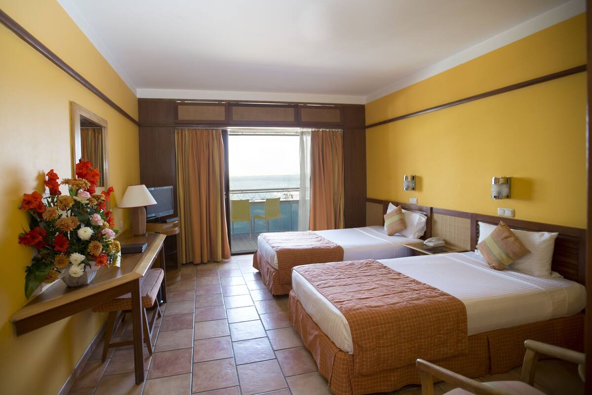Une chambre d’hôtel avec deux lits, un bureau avec une lampe, un vase de fleurs et une vue sur la mer par la fenêtre.