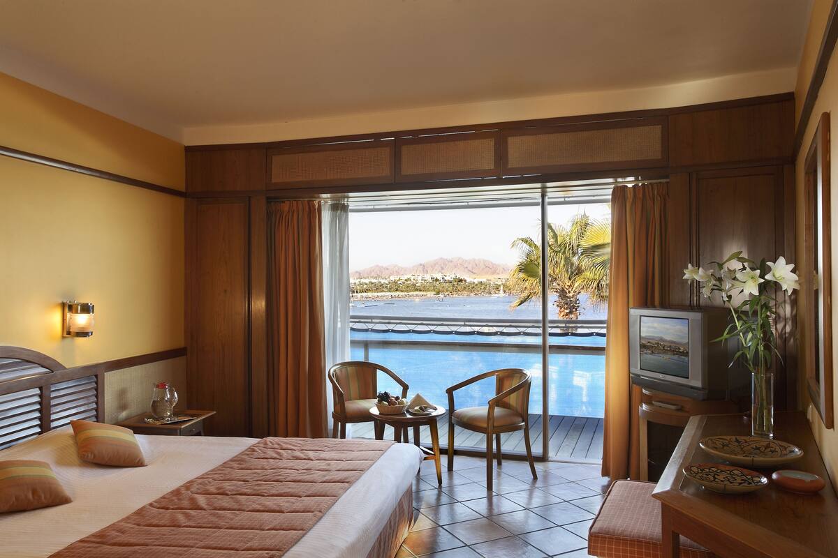 Une chambre d’hôtel avec un lit, deux chaises, une petite table ornée d’un vase de fleurs, un téléviseur et une vue sur une piscine et des montagnes.
