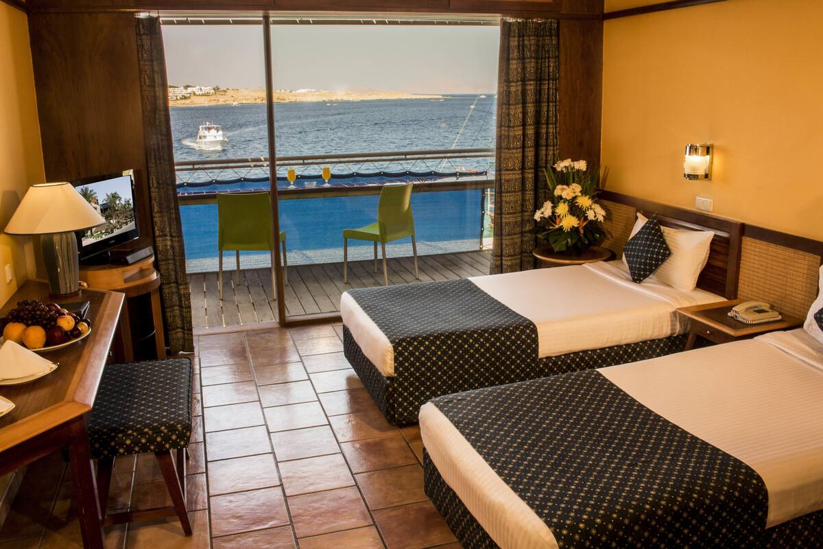 Une chambre d’hôtel avec deux lits, un bureau avec une lampe, une chaise, une petite table avec un vase de fleurs, et une vue sur la mer et un bateau.
