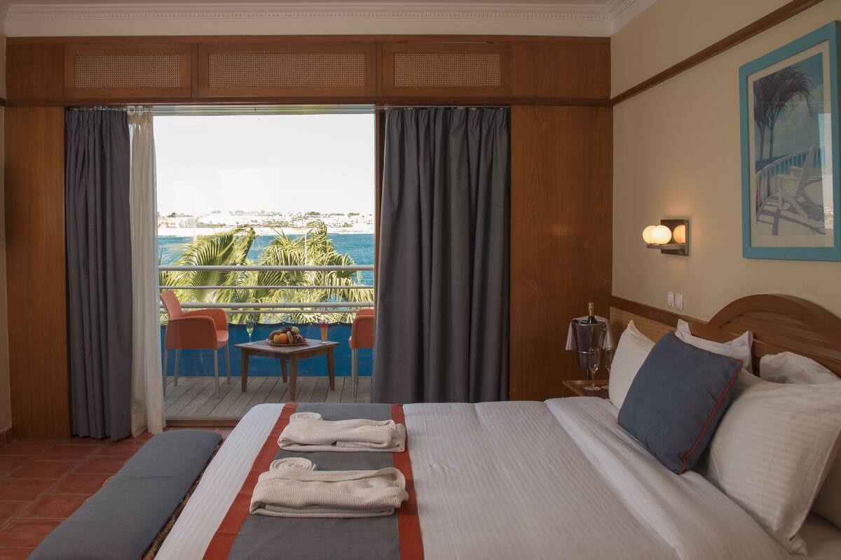 Une chambre d’hôtel avec un lit, une télévision, un balcon avec une table et des chaises, et une vue sur la mer.