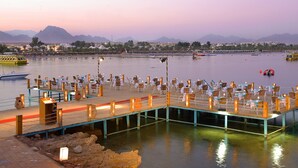 Un restaurante frente al agua con terraza y vistas al mar, un muelle con barcos y montañas al fondo.