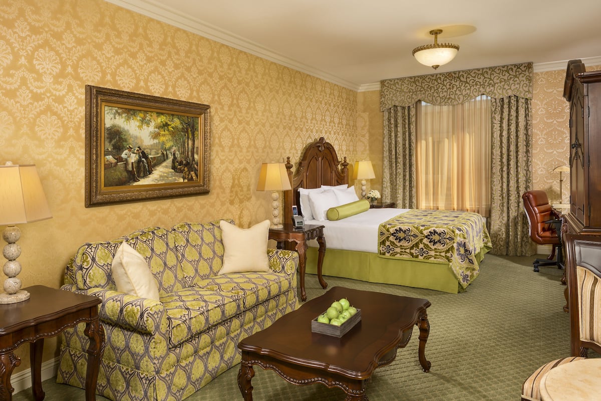 Een hotelkamer met een gestreept bankje, een bed met groene beddengoed, een houten bijzettafel met appels, en een schilderij aan de muur.