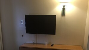 A hotel room with a flat-screen TV mounted on the wall, a wooden dresser with drawers, a remote control on the dresser, and a small wall-mounted light fixture. - Caravan Motel (Niagara Falls)