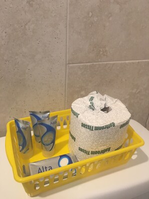 Keltainen kori, jossa on wc-paperia, kaksi pakettia Alta-shampoota ja wc-paperirulla, jonka kuosissa on sarjakuvahahmo.