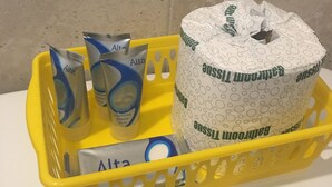 A yellow basket containing toiletries including two packets of shampoo, a bottle of conditioner, and a roll of toilet paper. - Caravan Motel (Niagara Falls)