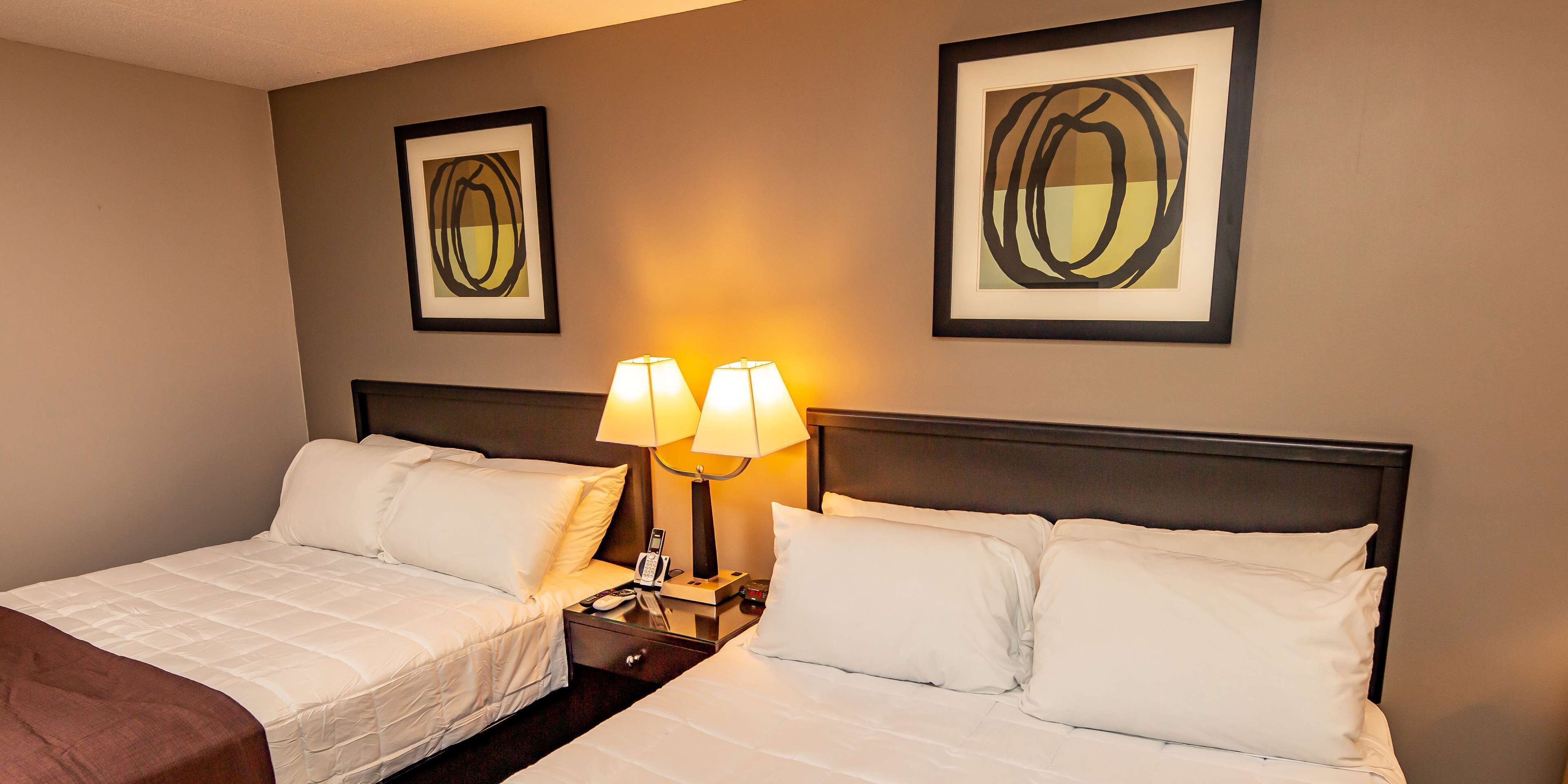 A hotel room with two beds, a nightstand with lamps, and two framed abstract paintings on the wall.