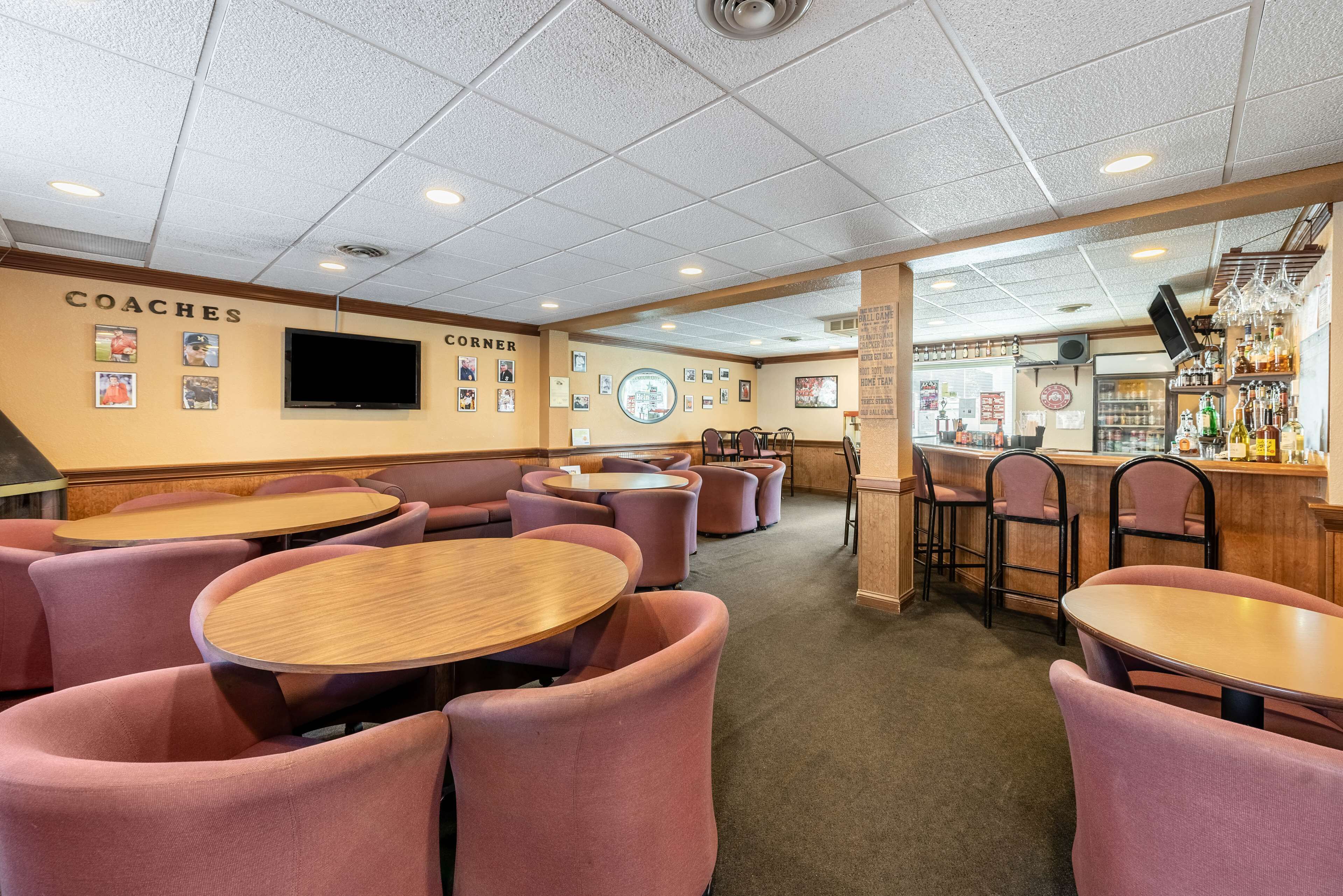 Un restaurante con mesas redondas y sillas acolchadas, una zona de bar con taburetes, y una pared decorada con imágenes y la palabra 'COACHES'.