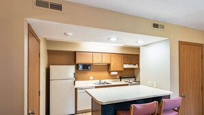 A compact kitchen with wooden cabinets, a white refrigerator, a microwave, a stove, and a sink. There are two pink chairs at the island.