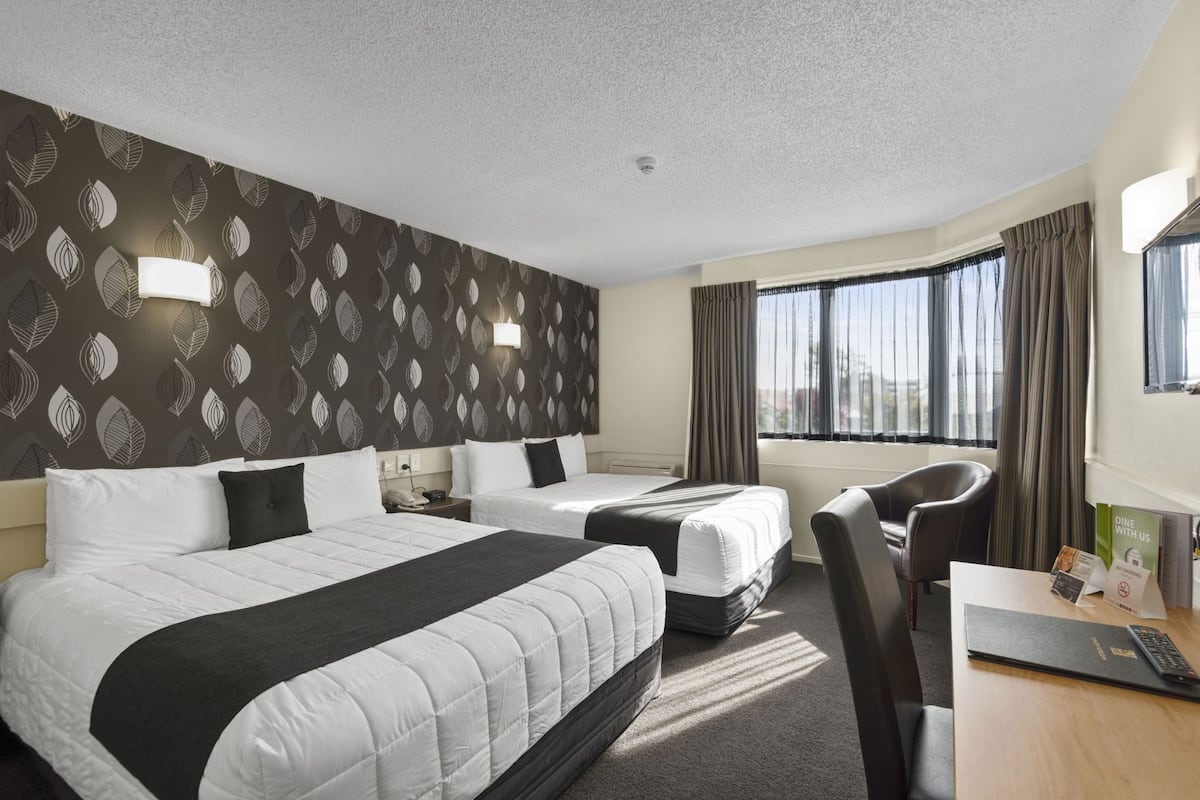 Camera d'hotel con due letti, una scrivania, una sedia, una finestra con tende e un muro decorato.