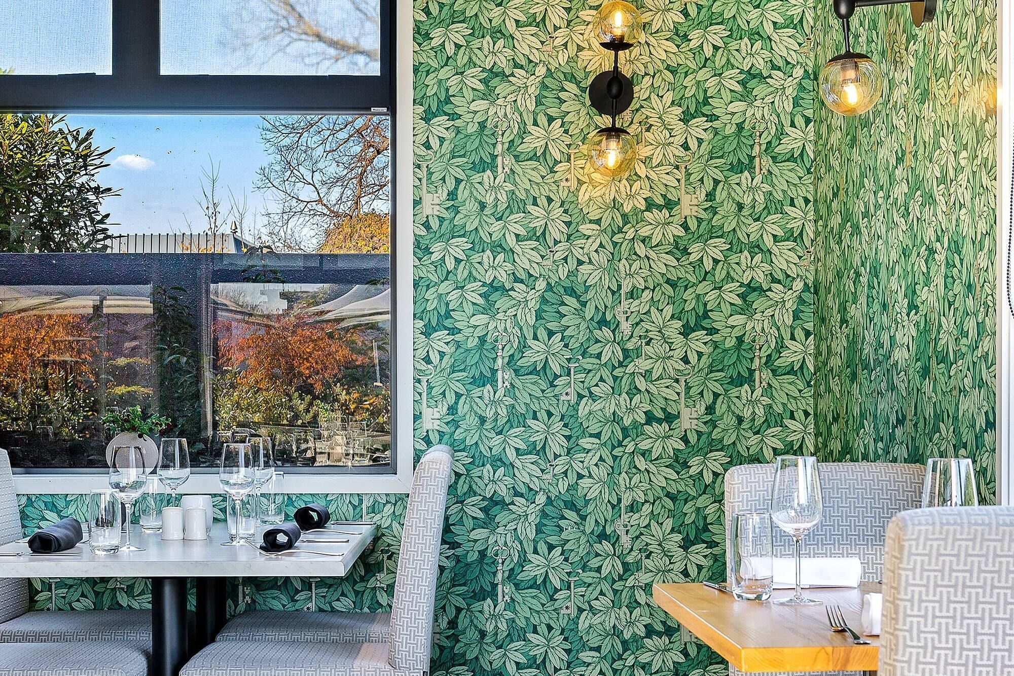 Ein Restaurant mit Blick durch ein Fenster, Tische mit Weingläsern und einer grünen, blattigen Wand.